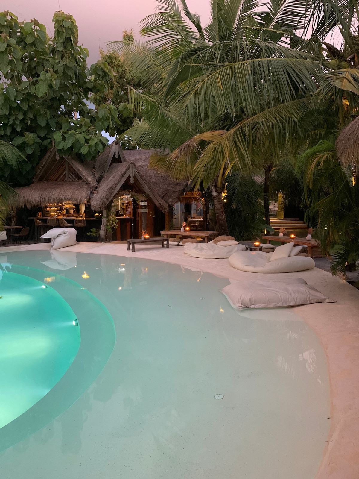 A tranquil pool area is framed by lush palm trees and cozy seating. Soft cushions are scattered around the pool's edge, providing a relaxed atmosphere. The gentle lighting reflects off the water, enhancing the inviting feel of this outdoor space.