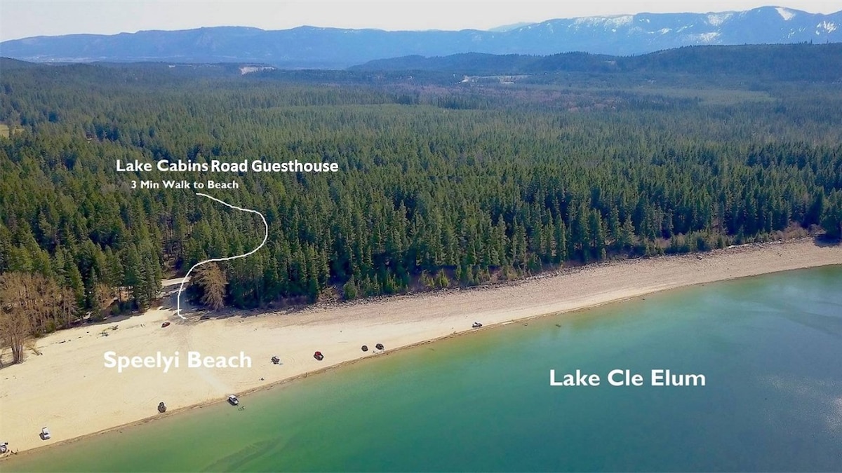 An aerial view captures the proximity of the Lake Cabins Road Guesthouse to Speelyi Beach and Lake Cle Elum. Lush green trees frame the shoreline, with a sandy beach stretching along the tranquil waters. The designated walking path to the beach is highlighted, conveying easy access.