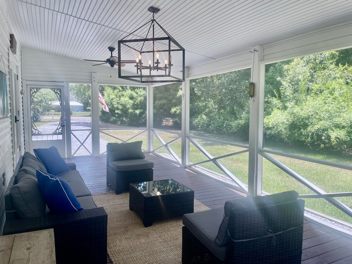 A spacious screened-in porch features comfortable seating arrangements, including a dark wicker sofa with blue cushions, and a glass-top coffee table. Soft natural light fills the area, surrounded by lush greenery visible through the screens. A ceiling fan provides a cooling breeze.
