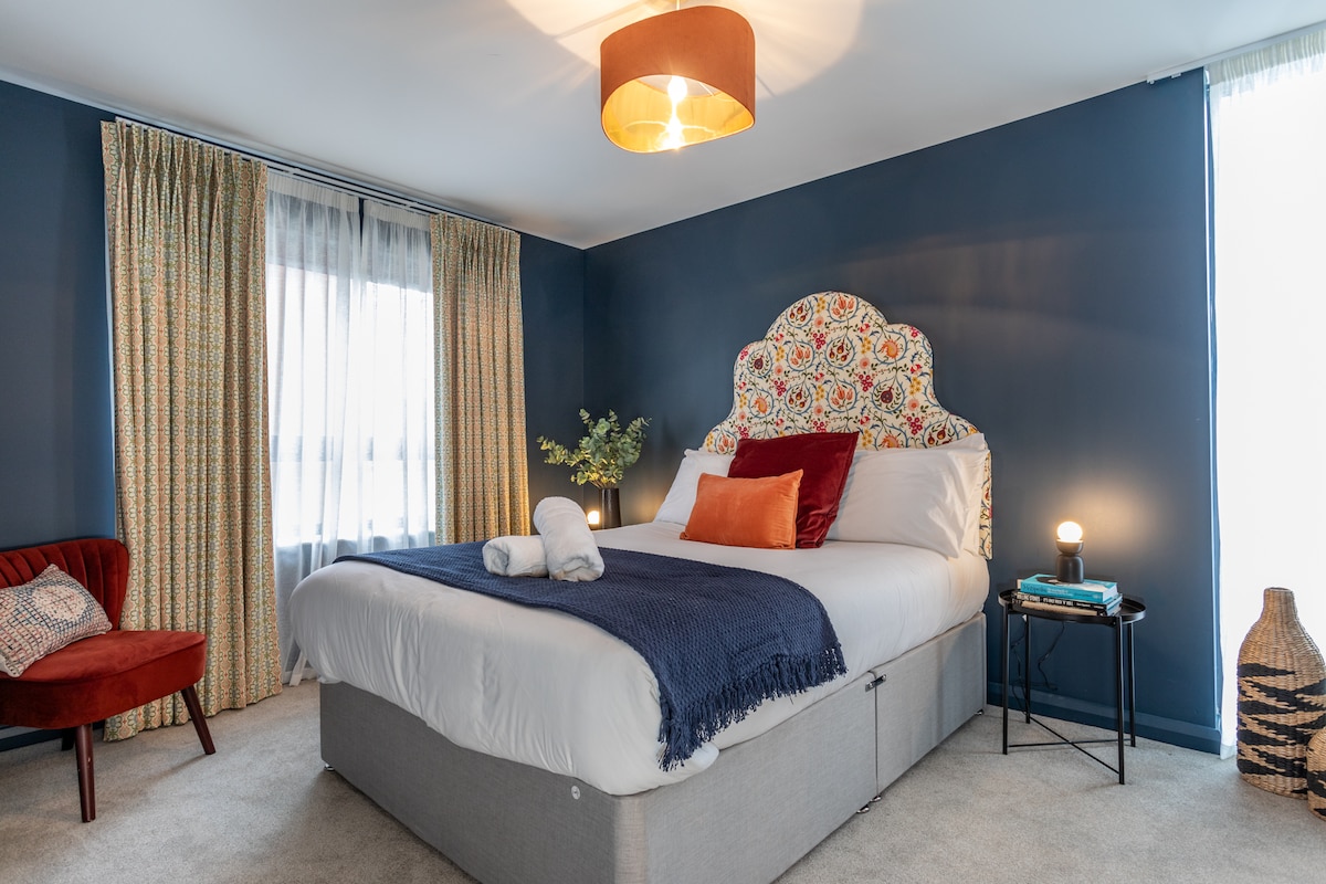 A bedroom features a spacious bed with a decorative headboard, complemented by colorful accent pillows. A cozy chair and side table are positioned nearby. Soft curtains frame the windows, allowing natural light to brighten the room, while a pendant light adds a warm glow.