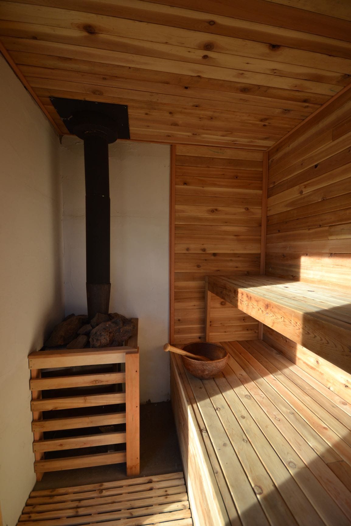 A wood-fired sauna is presented with natural wood paneling, creating a warm and relaxing environment. A metal stove is positioned in the corner, alongside a wooden bucket and ladle. The two-tiered seating ensures ample space for guests to unwind.