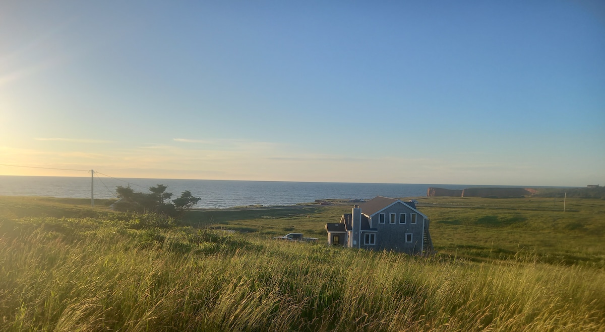 A coastal view captures a serene home set against a backdrop of open grassland and the sea. The water sparkles under a clear sky as soft sunlight illuminates the scene, highlighting the natural beauty of the surroundings.