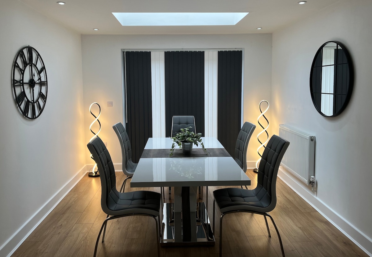 A modern dining area features a sleek table surrounded by six contemporary chairs. Natural light is enhanced by a skylight and accentuated by wall-mounted mirrors and decorative standing lamps. Dark curtains frame the window, creating a balanced and inviting environment.