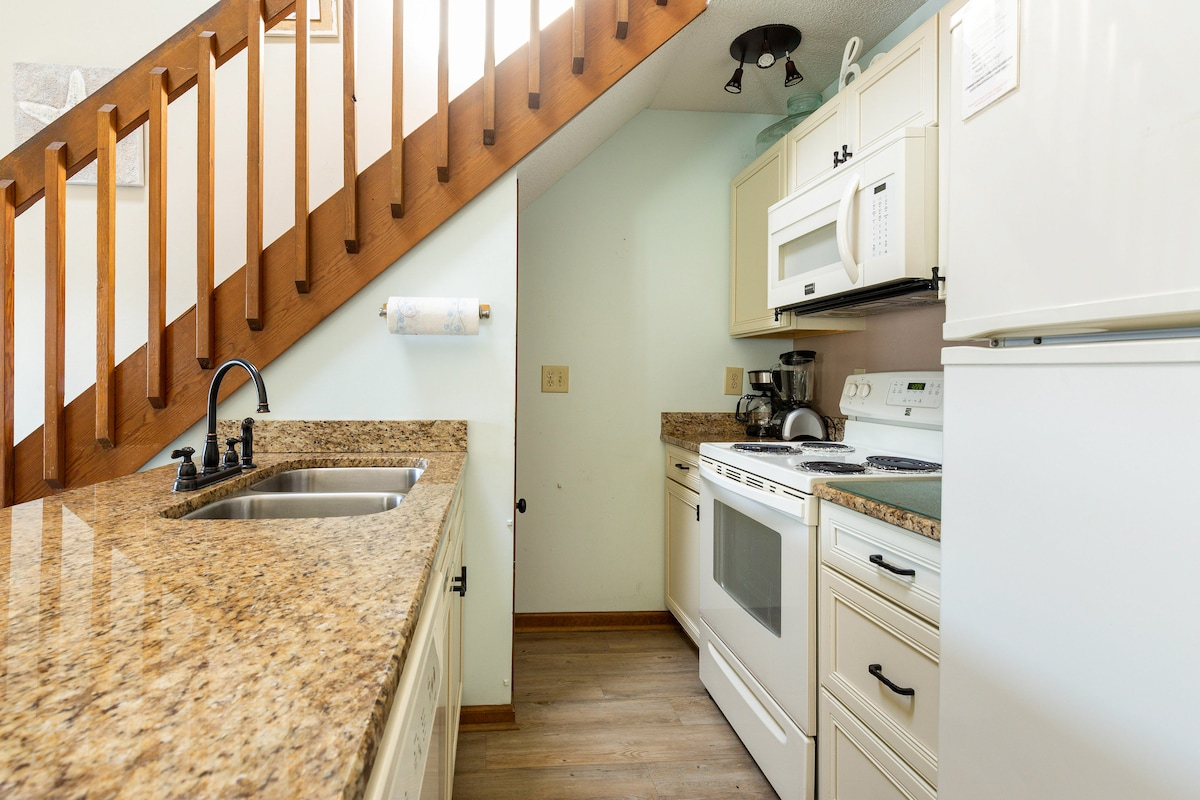 A fully-equipped kitchen showcases granite countertops and white cabinetry. Appliances include an oven, stove, microwave, and refrigerator. A sink is positioned below a window, and a coffee maker is visible. Stairs extend above, with wooden railing adding warmth to the space.