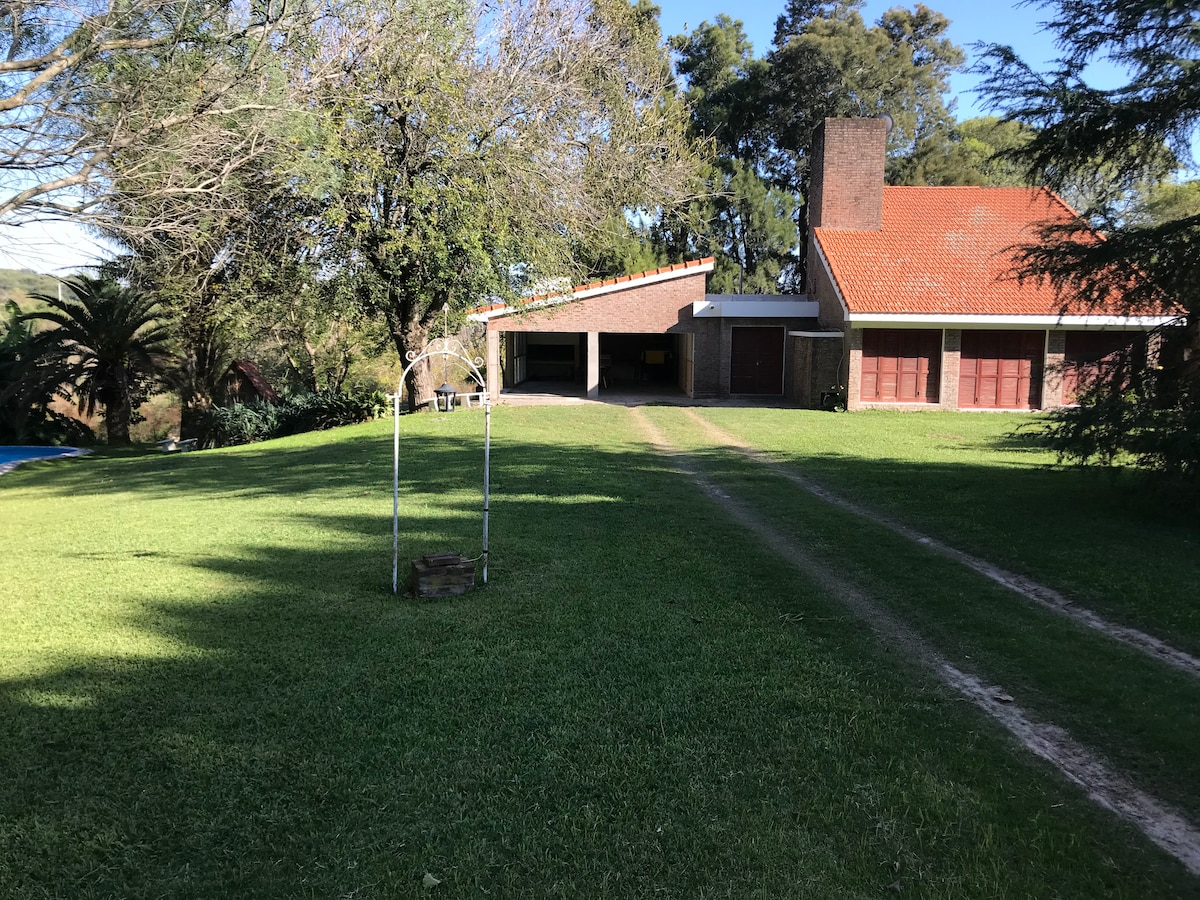A lush green lawn stretches out towards a red-roofed house, showcasing a blend of brick and wood elements. Large trees provide shade, while a pathway leads to the entrance. An archway stands on the lawn, hinting at outdoor recreation opportunities.