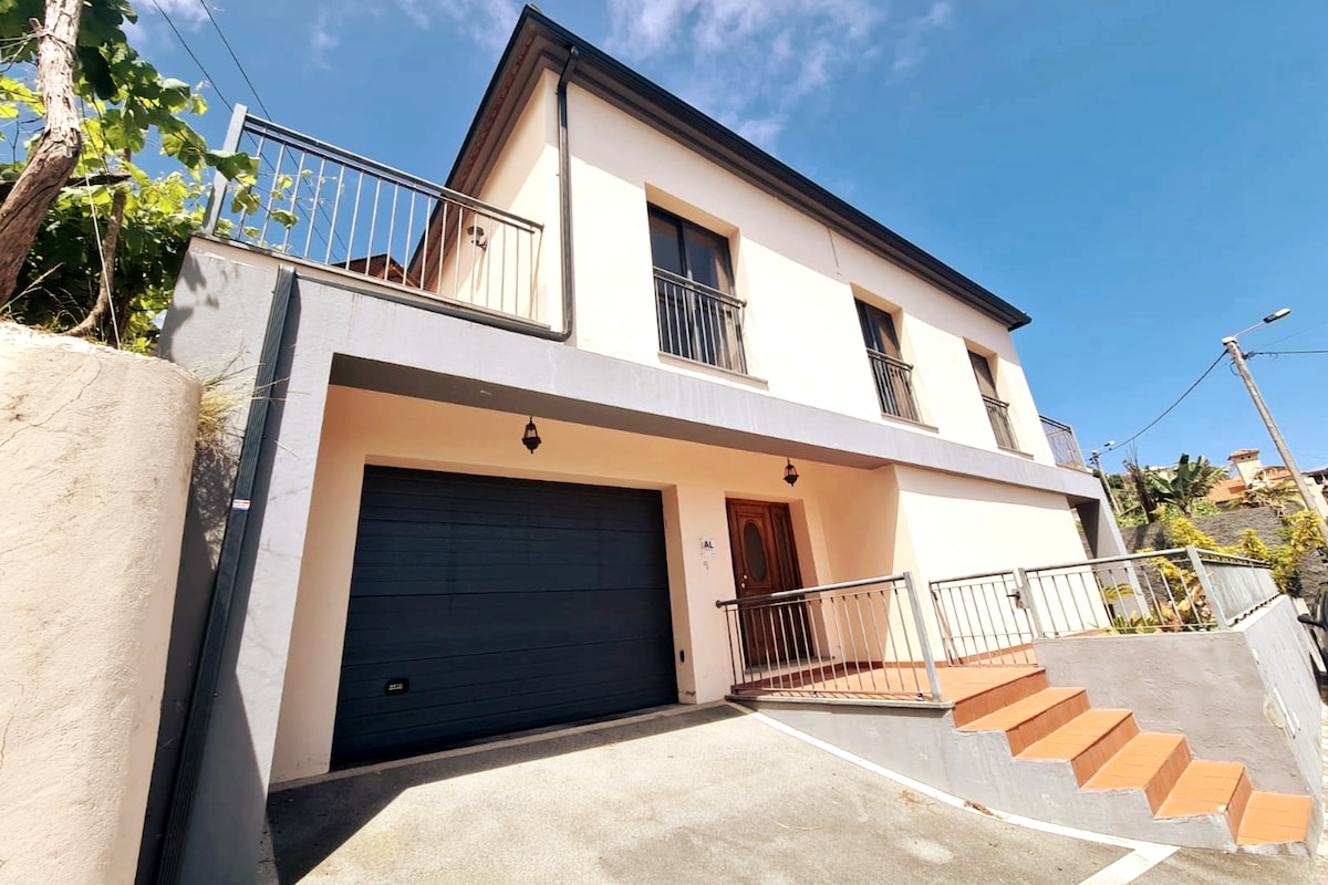 A modern two-story house with a light-colored façade is shown. Steps lead up to the entrance, framed by a small garden. A garage door is visible on the left, and a balcony with black railings is situated above the entrance, offering a view of the surrounding area.