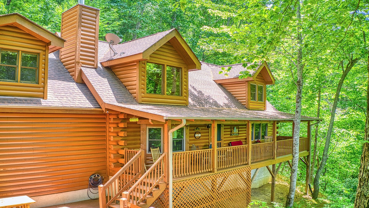 The exterior of a spacious log cabin is surrounded by lush greenery, featuring a covered porch with wooden railings and a set of stairs leading down. Large windows allow for natural light, enhancing the inviting appearance of the home.
