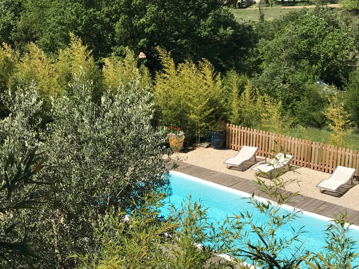 A communal swimming pool is set within a lush garden featuring various trees and plants. Sun loungers are positioned along the pool's edge, which is bordered by a wooden deck, providing a calm atmosphere amid natural surroundings.