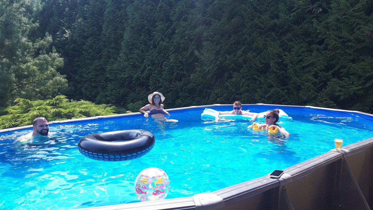 An above-ground pool is surrounded by lush greenery, offering a refreshing blue space for relaxation. Inflatable floats are present, and several individuals are enjoying the water. Sunlight reflects off the surface, enhancing the tranquil atmosphere.
