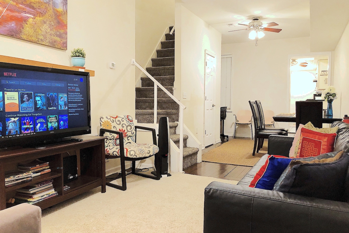 The living area features a dark sofa accented with vibrant cushions, a flat-screen television displaying streaming options, and a cozy carpet. A dining space with dark chairs is visible, accompanied by a staircase leading to the upper levels of the townhouse.