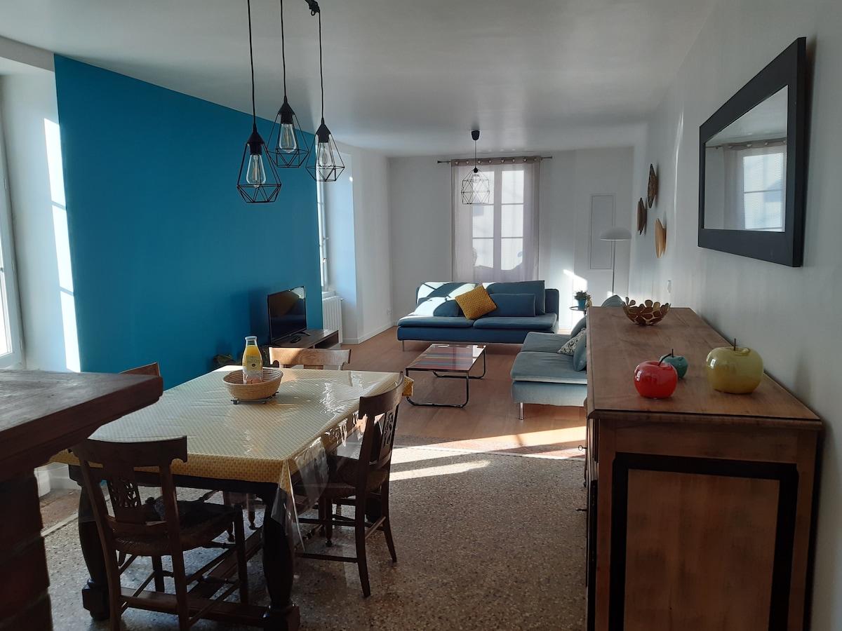 A spacious living area is presented with a blue accent wall, complemented by natural light from large windows. A comfortable sofa sits opposite a coffee table, while a dining table is surrounded by wooden chairs. Decorative elements include a bowl of fruit on the sideboard.