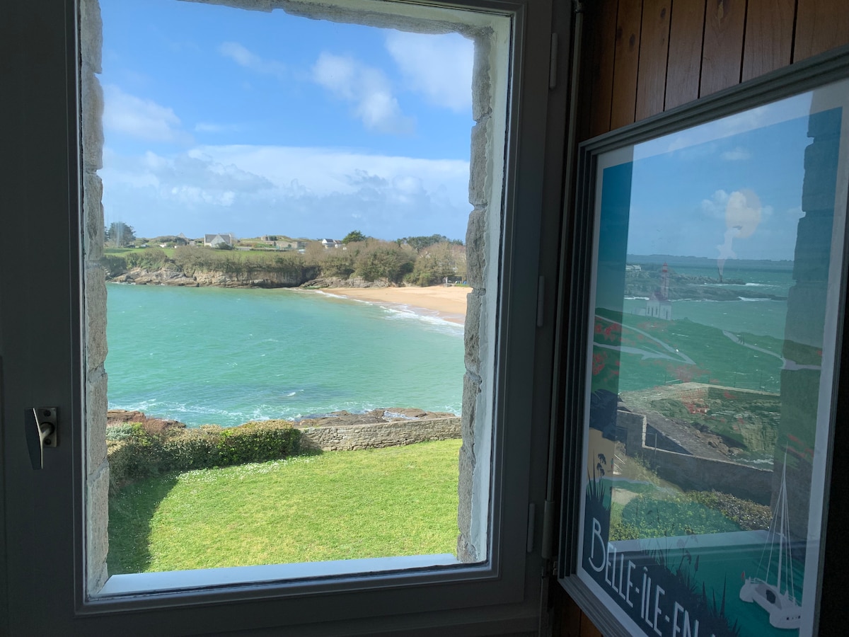 A window provides a view of the vibrant turquoise water and sandy beach, with green hills visible in the distance. The landscape features lush grass and coastal vegetation. The sky is partly cloudy, enhancing the coastal scenery.