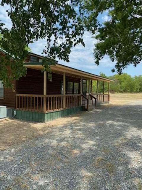 The exterior of a wooden cabin is shown, featuring a covered porch that spans the front. Steps lead up to the entrance, surrounded by a gravel driveway. Trees provide partial shade, and a spacious, natural area extends beyond the cabin.