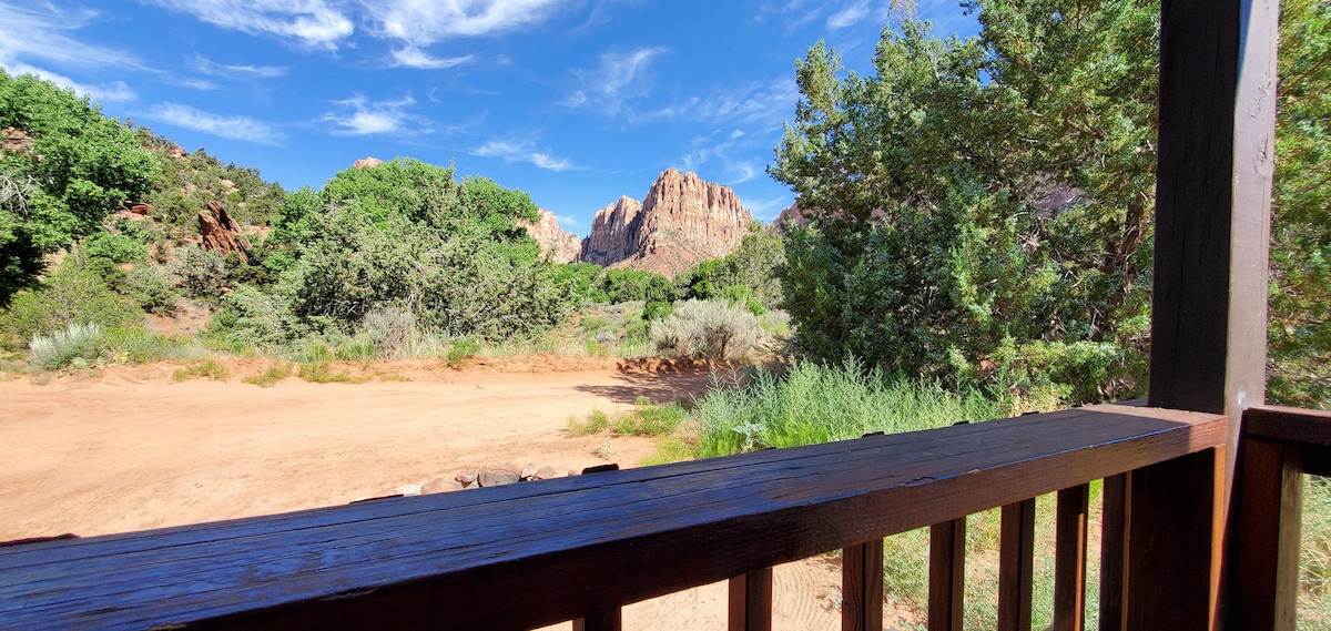 A wooden railing frames a scenic view of the landscape beyond. Lush greenery contrasts with the reddish-brown mountain formations under a clear blue sky, highlighting the natural beauty of the surrounding canyon area.