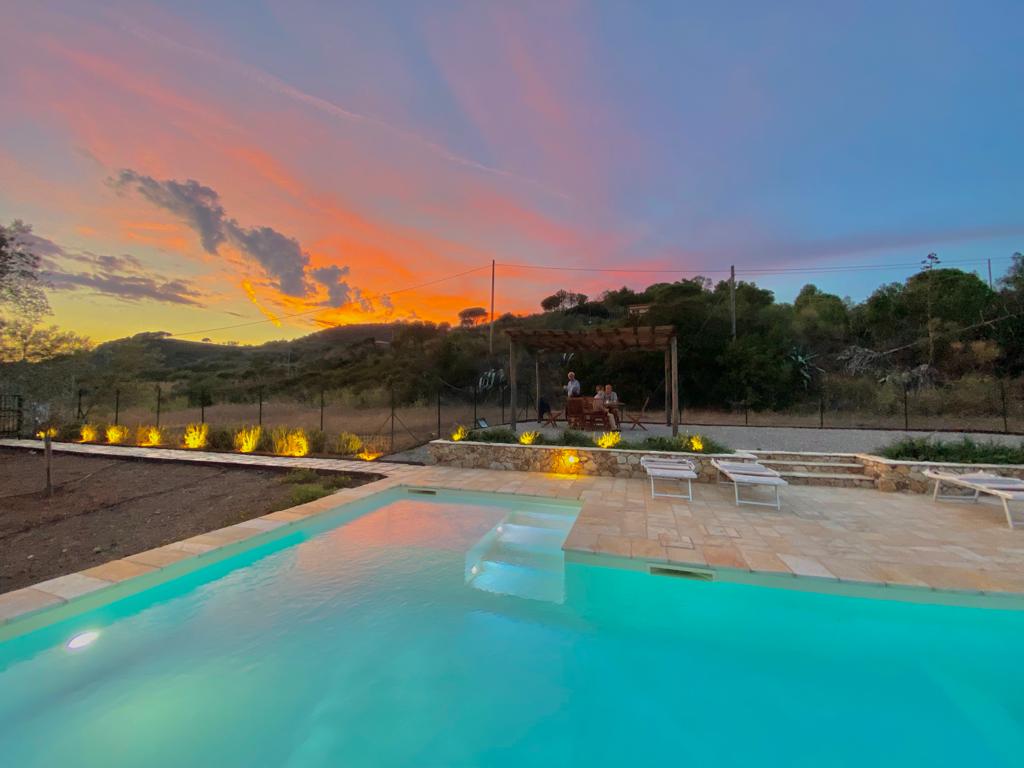 A serene pool area is framed by a soft landscape, showcasing calm turquoise water reflecting a vibrant sunset sky. Lounge chairs are positioned alongside the pool, with soft lighting illuminating the surrounding garden. A rustic pergola can be seen in the background, enhancing the tranquil atmosphere.