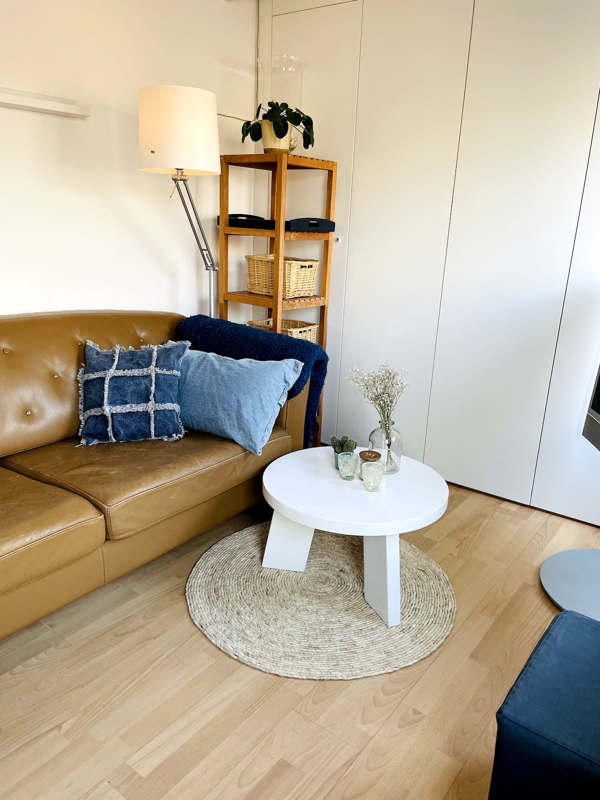 A cozy living space contains a brown leather sofa adorned with decorative pillows and a navy throw. A small round white table is positioned in front, complemented by a woven rug. Natural light enters through a nearby window, enhancing the inviting atmosphere.