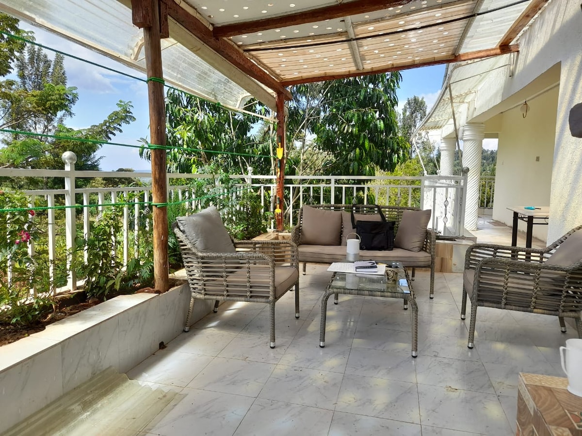 A cozy outdoor seating area is featured with wicker chairs and a glass table under a shaded patio roof. Lush greenery surrounds the space, providing a tranquil environment. Natural light filters in, highlighted by the bright blue sky above.