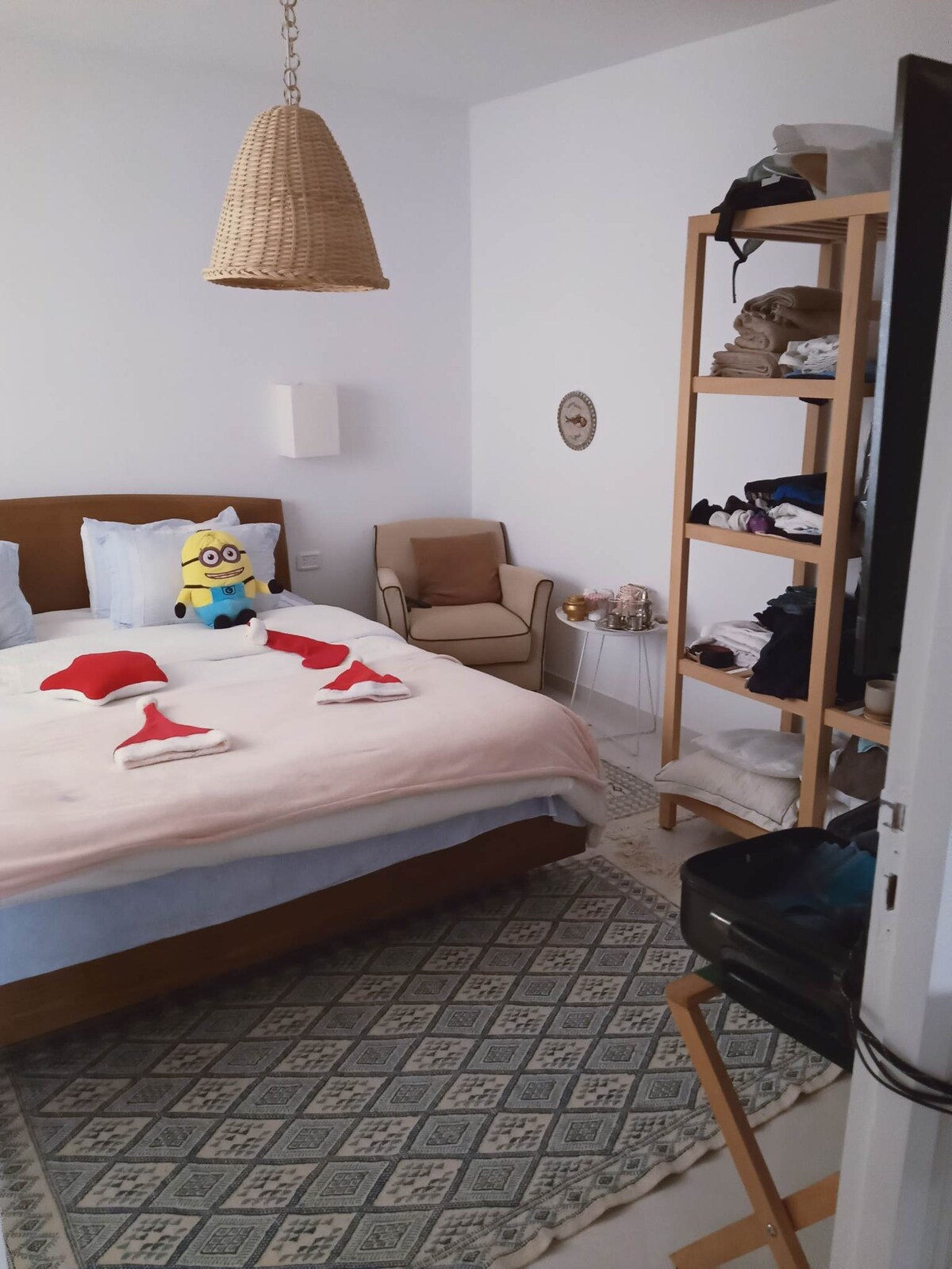 A tastefully arranged bedroom features a spacious bed adorned with creative towel arrangements. A plush character sits at the head of the bed. A wooden shelving unit displays neatly folded linens. A cozy chair is positioned nearby, complemented by a patterned rug underfoot.