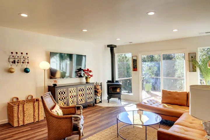 A well-lit living area is presented with a large flat-screen TV mounted on the wall and a cozy wood stove. A comfortable leather sectional and a round coffee table complement the natural decor. Large glass doors provide access to the outdoor space, enhancing the connection with nature.