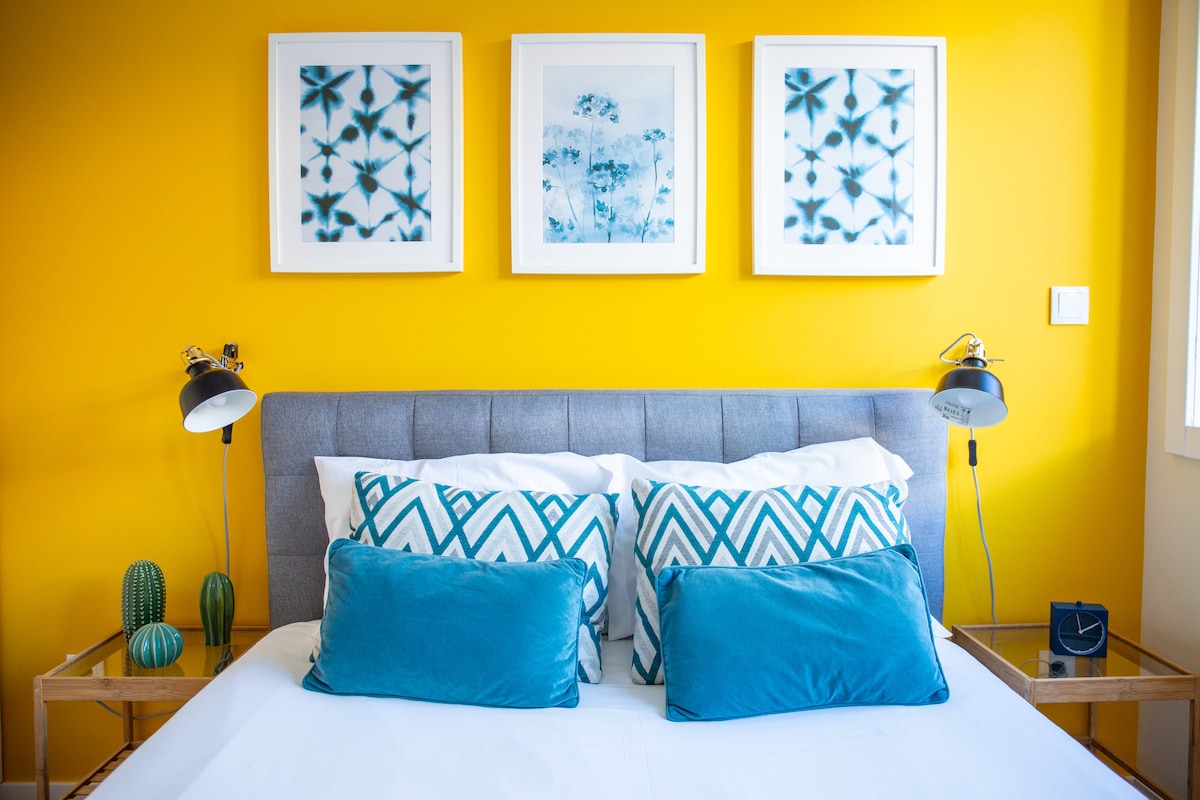 A modern bedroom features a gray upholstered headboard against a bright yellow wall, accented by three framed blue and white artworks. Two decorative pillows in shades of teal sit on the spacious bed, accompanied by stylish bedside lamps. Small potted cacti add a touch of greenery.
