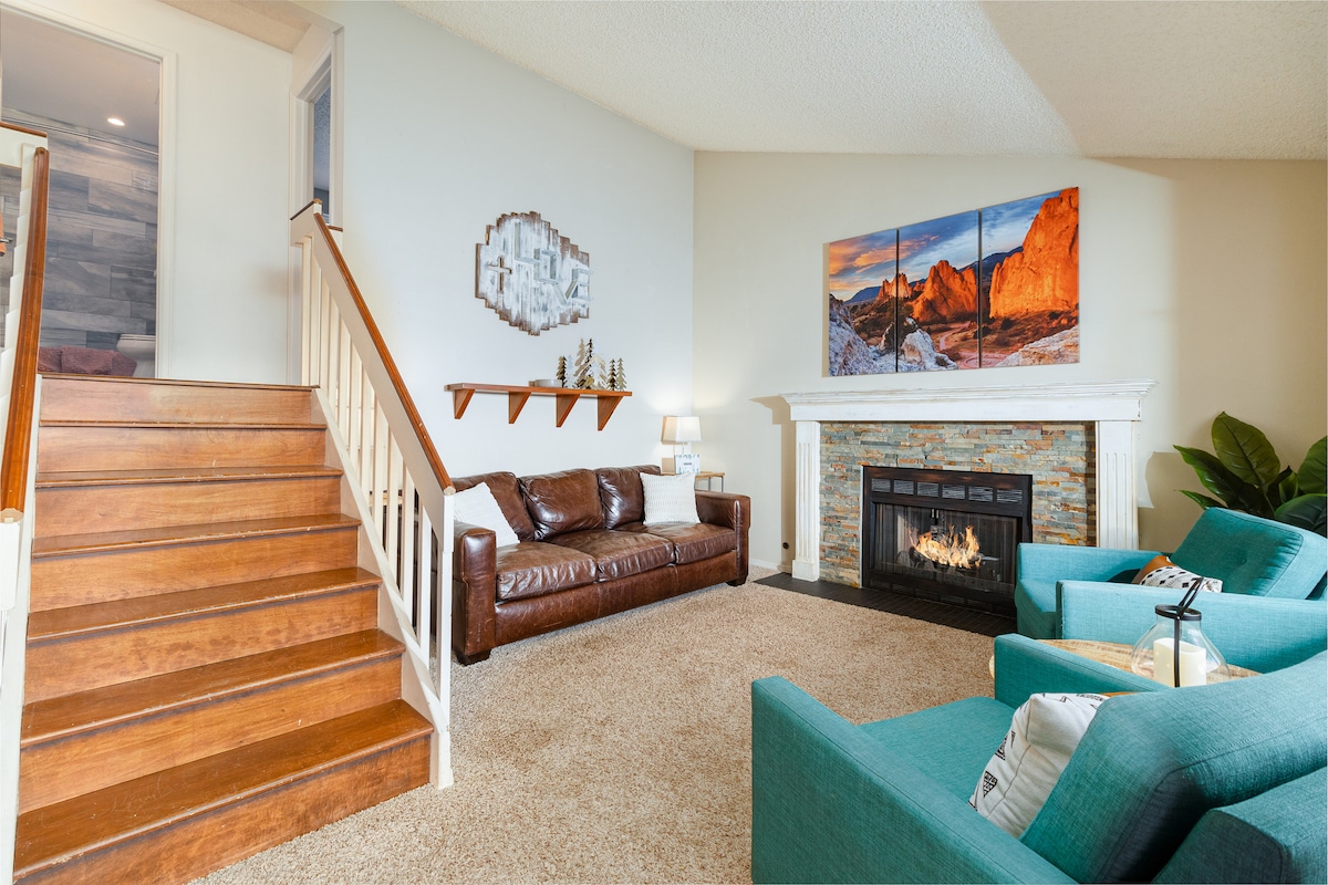 The living room features a cozy brown leather sofa and two turquoise armchairs, arranged around a stone fireplace. A large landscape painting hangs above the mantel, and a staircase leads to the upper level. Soft carpeting enhances the room's warmth.