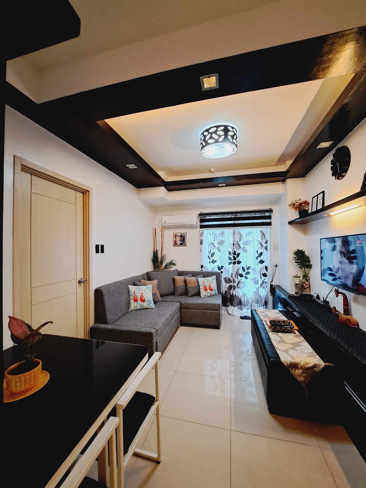 The living area features a modern grey sofa arranged around a sleek center table. A flat-screen television is mounted on the wall, complemented by decorative shelves. Natural light is encouraged through the floor-to-ceiling window, adorned with sheer curtains, while cove lighting enhances the overall space.