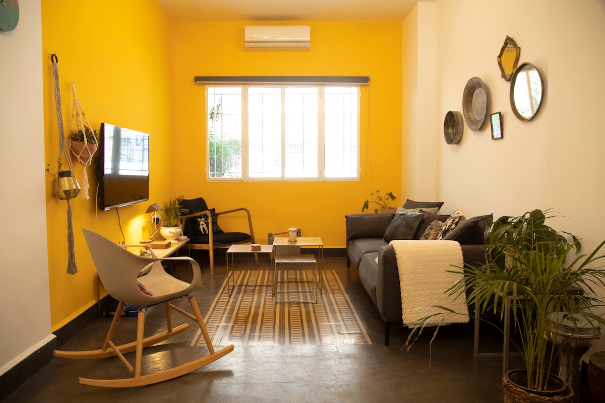 The living area features a bright yellow wall and a mix of modern and antique decor. A comfortable seating arrangement includes a sofa and a rocking chair, positioned around a low coffee table. Natural light floods in through large windows, enhancing the homey ambiance.