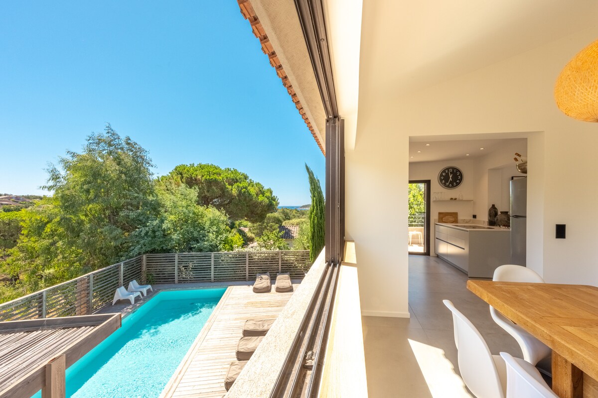 A view from the terrace showcases a refreshing pool surrounded by wooden decking and sun loungers. Lush greenery frames the area, while an open living space with a clock and a modern kitchen is visible indoors, offering a seamless connection between indoor and outdoor settings.