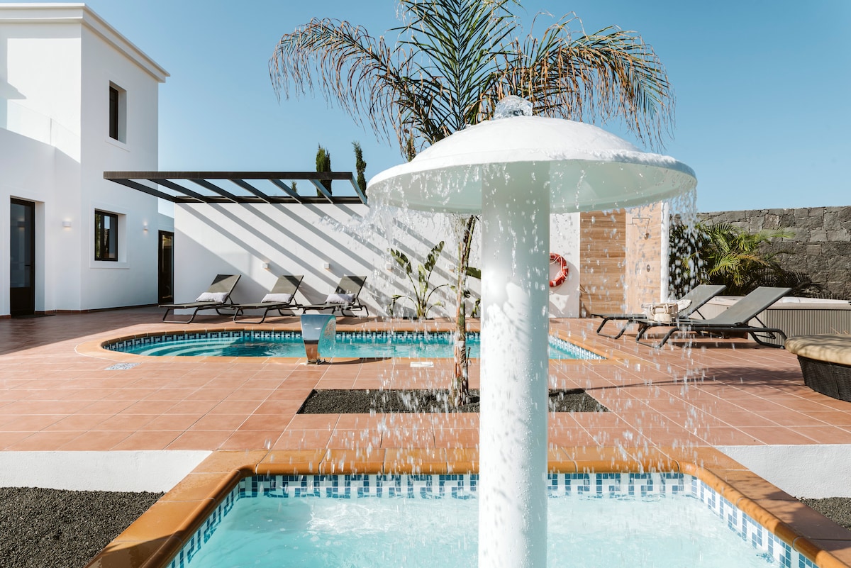 A crystal-clear fountain features prominently in the foreground, with water cascading gracefully. The surrounding area includes two pools, one for children, and several lounge chairs under a shaded area. Bright sunshine reflects off the tiled patio, enhancing the inviting atmosphere.