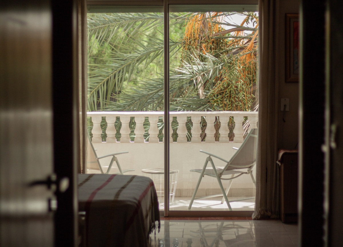 A large sliding glass door provides a view of a private balcony adorned with two white chairs. Lush palm tree leaves are visible, creating a serene outdoor setting. The softly lit interior includes a spacious bed and neutral-colored decor.