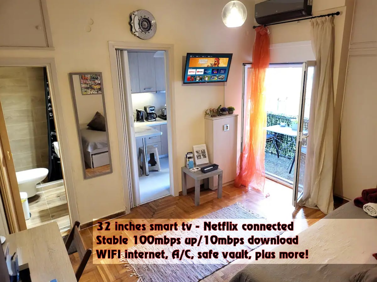 A compact living space is displayed, featuring a 32-inch smart TV connected to Netflix. Nearby, a fully equipped kitchen is visible, and a cozy sleeping area is shown with a soft bed covering. Natural light enters through a window leading to a balcony.