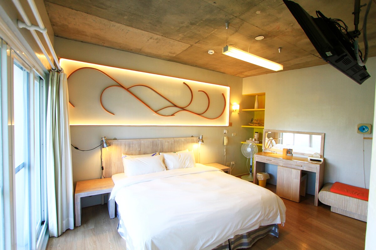 A bright room features a spacious bed with crisp white linens. Natural light filters through large glass doors, complemented by a modern wooden accent wall. A desk and chair occupy one side, with a ceiling-mounted television providing entertainment options.