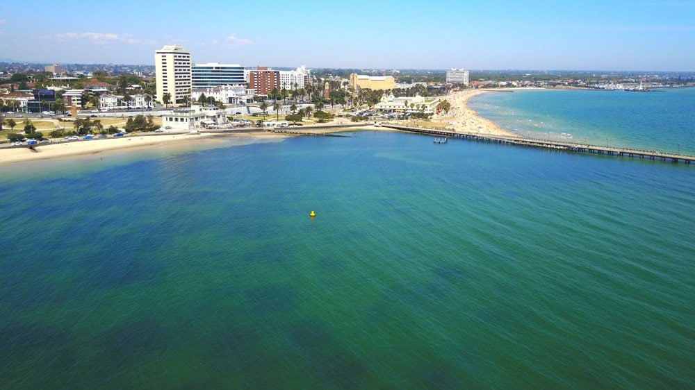An aerial view captures the serene bay with calm, green waters meeting a sandy beach. Lined with palm trees, a vibrant promenade and several nearby buildings can be seen, creating a coastal atmosphere. The horizon features a clear blue sky dotted with a few clouds.