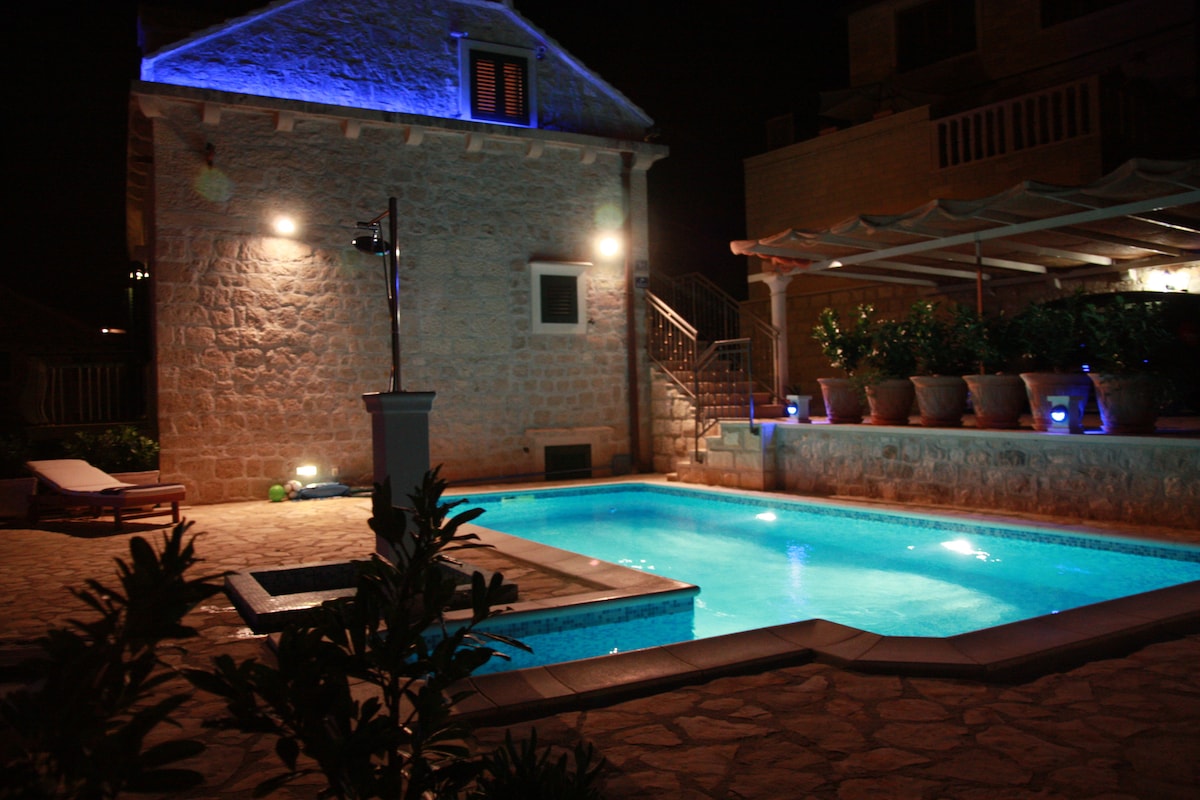 A tranquil night scene showcases a stone house illuminated by soft blue lighting. The inviting swimming pool, surrounded by a tiled patio, reflects the glow. Nearby, a covered terrace features potted plants, enhancing the outdoor living space.