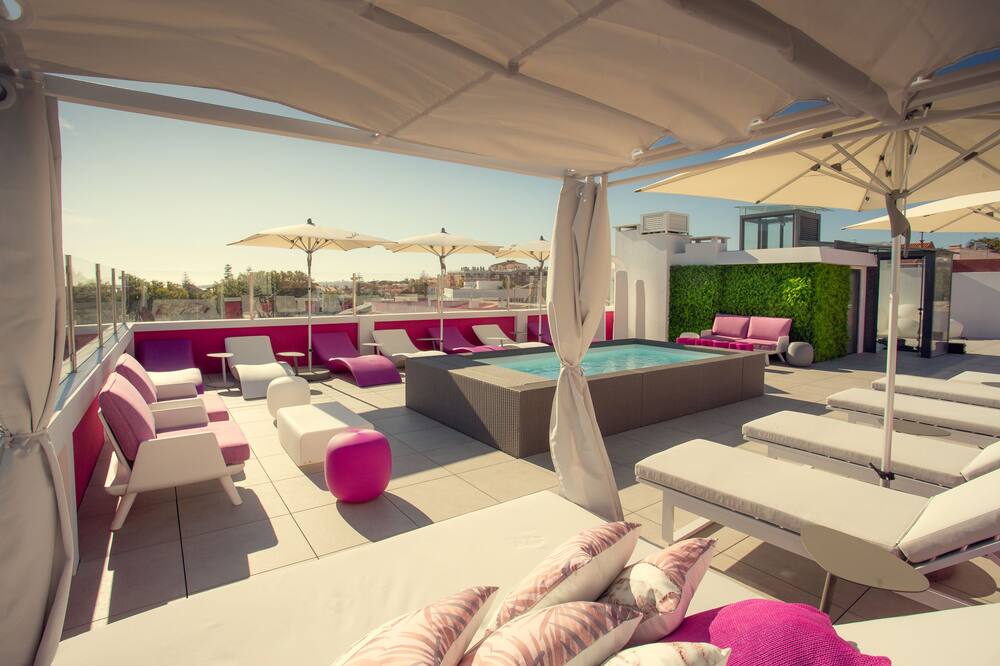 The rooftop area features a small pool surrounded by sun loungers and shaded cabanas. Bright pink accents complement the neutral tones of the seating. Umbrellas offer shade, creating a welcoming space for relaxation with views of the surrounding area.
