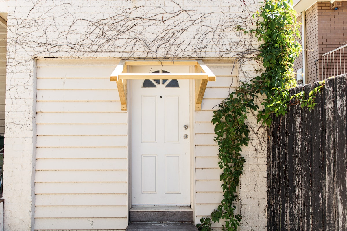 A private entrance is featured, set within a light-colored wooden exterior. A small roof overhang extends over the door, which is complemented by climbing greenery along the side. The entrance is framed by a rustic wooden fence on one side.