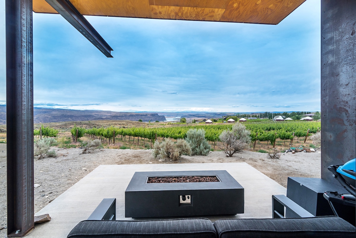 An outdoor seating area features a modern fire table surrounded by comfortable seating. The expansive view showcases rolling hills adorned with greenery, creating a serene backdrop. Under a partially covered patio, the scene is complemented by distant vineyards and soft, cloudy skies.