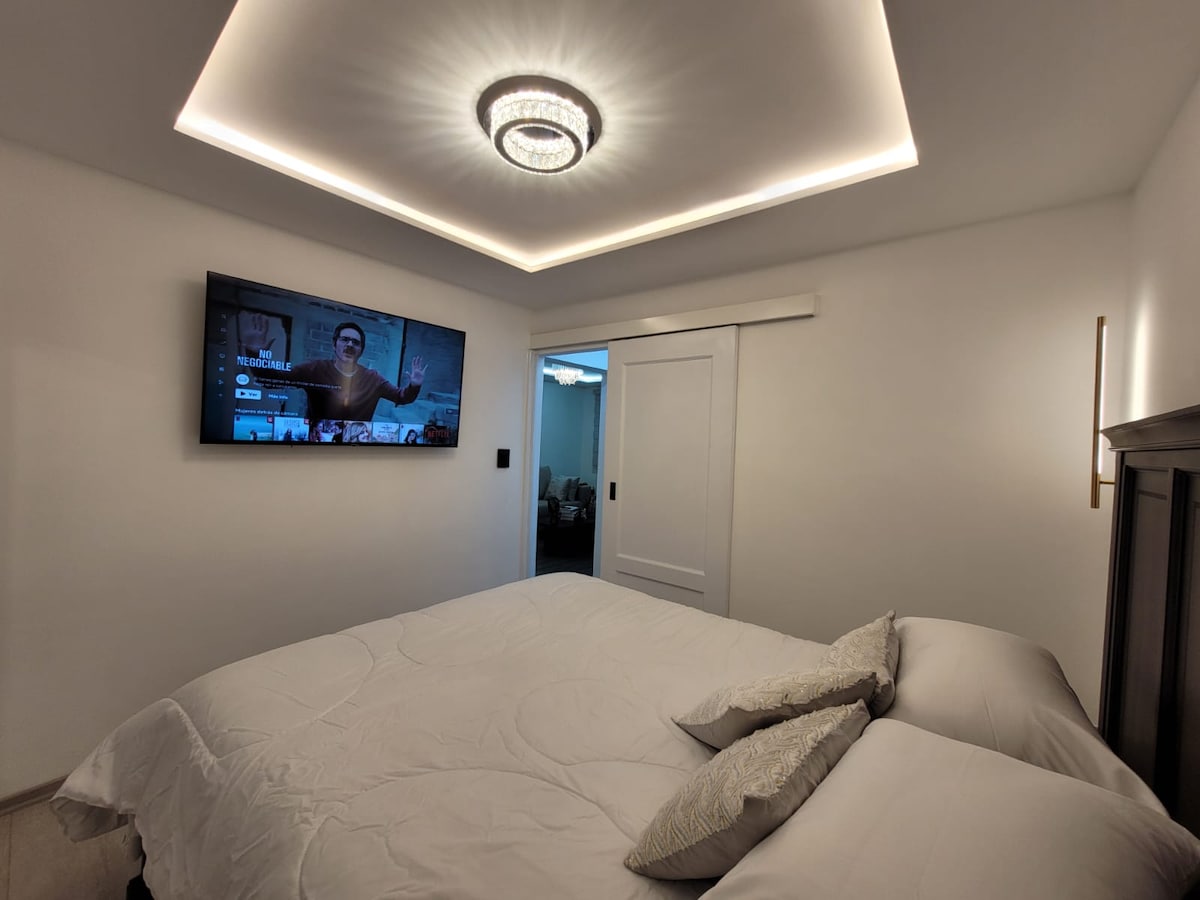 A modern sleeping area is presented with a spacious bed featuring light-colored linens. A flat-screen television is mounted on a white wall, while soft lighting from the ceiling adds a contemporary touch. Sliding doors provide access to additional living spaces.