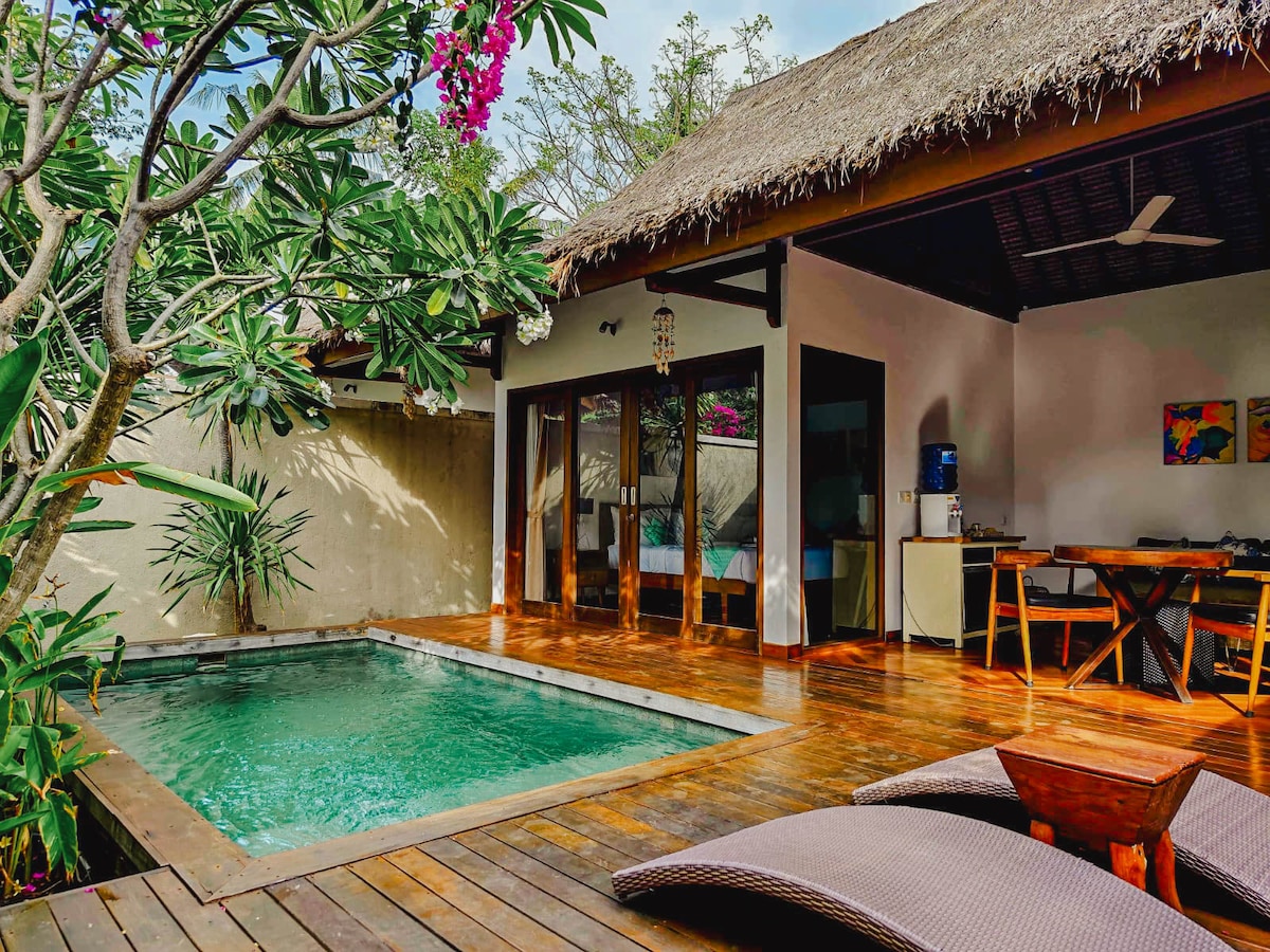 An outdoor area features a private swimming pool surrounded by a wooden deck and lush tropical plants. The villa's semi-open design includes large glass doors, allowing natural light to illuminate the space, which contains a dining table and seating area.
