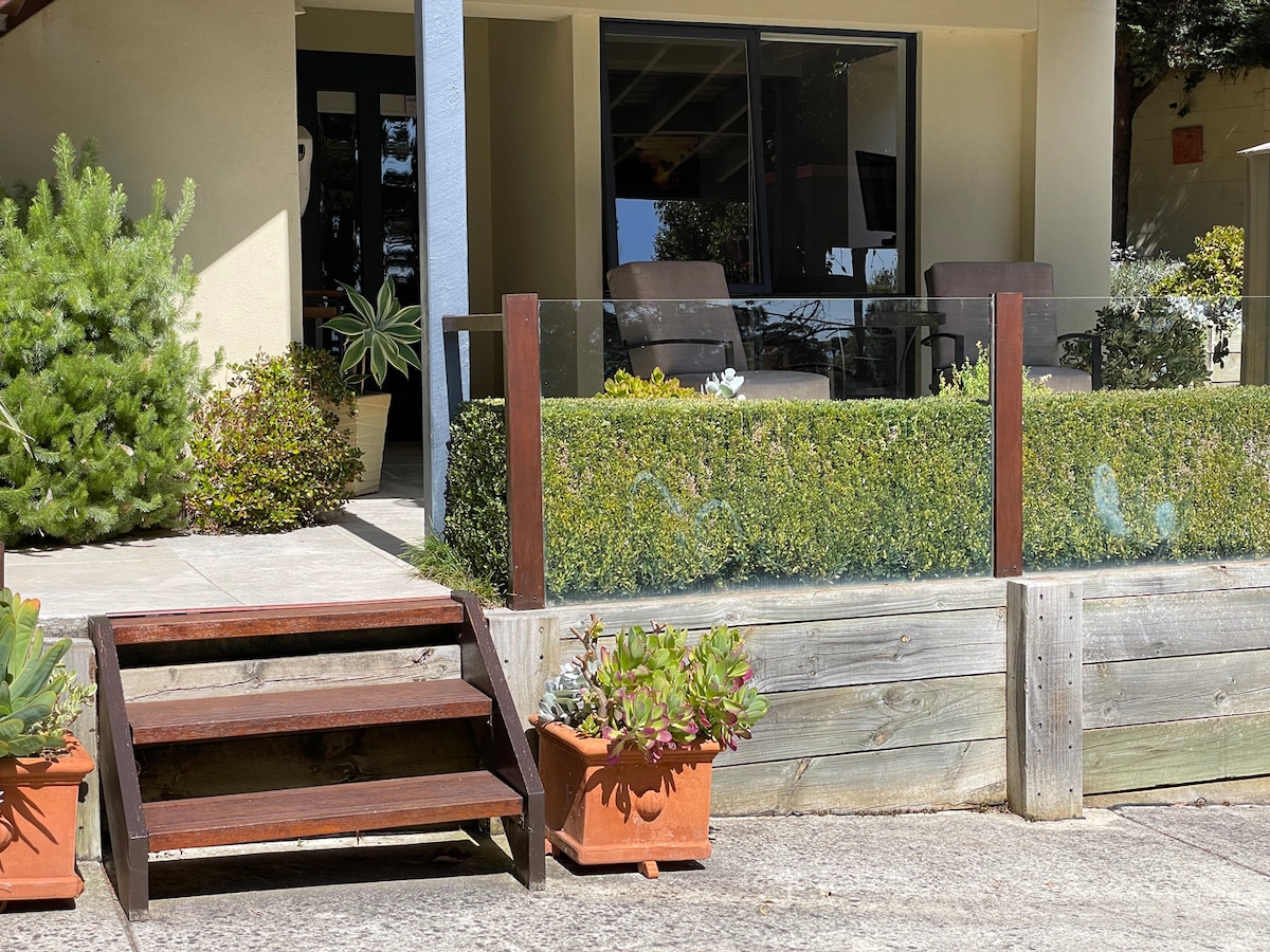 An outdoor area features a set of steps leading to a patio space surrounded by greenery. Two comfortable chairs are positioned behind a low glass barrier, allowing for unobstructed views of the garden. Potted plants add a touch of color to the setting.