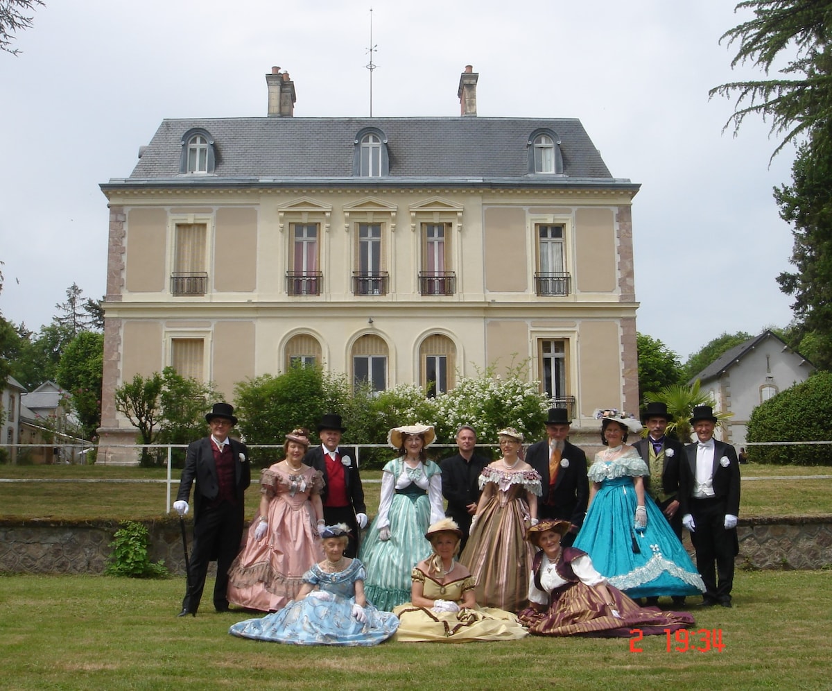 An elegant historical building is depicted in the background, showcasing a classic façade with symmetrical windows and a gray roof. In the foreground, a group of individuals dressed in vintage attire stands on a manicured lawn, surrounded by lush greenery and blooming bushes.
