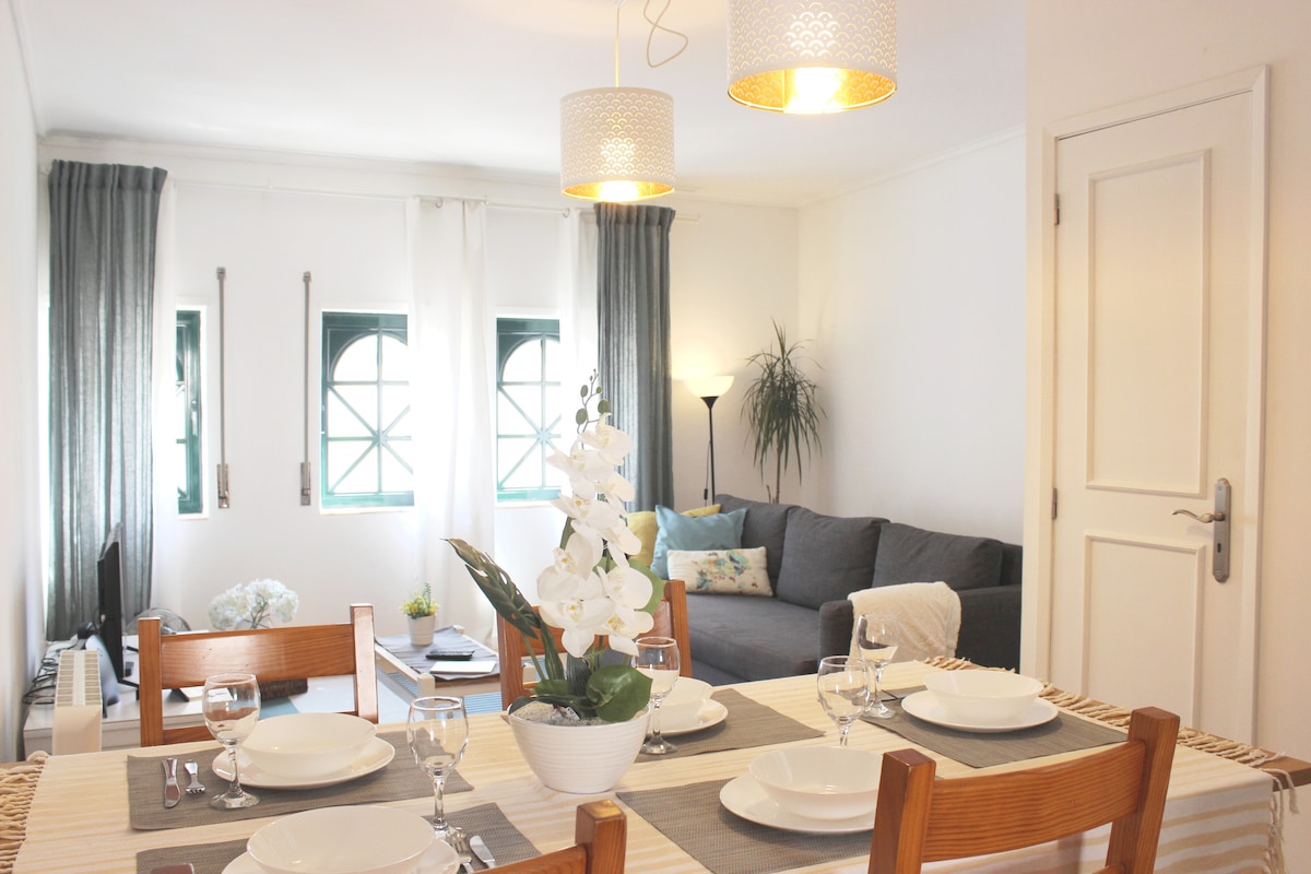 A dining area is featured, showcasing a wooden table set with plates and glasses. Two decorative pendant lights illuminate the space, complemented by a cozy seating area with a gray sofa. Natural light enters through large windows, enhancing the inviting ambiance.