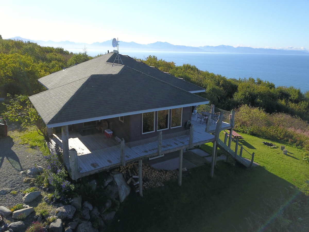 A custom-designed house with a unique circular roof sits on a bluff, surrounded by lush greenery. A spacious deck extends from the home, offering views of Kachemak Bay and the Alaskan Mountain range in the background.