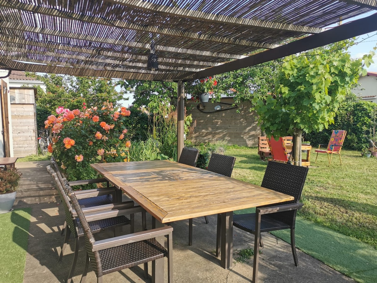 An outdoor dining area is shown, featuring a large wooden table surrounded by six wicker chairs. A pergola overhead offers shade, and vibrant roses bloom nearby, enhancing the natural setting. The spacious garden is visible in the background, highlighting the serene outdoor atmosphere.