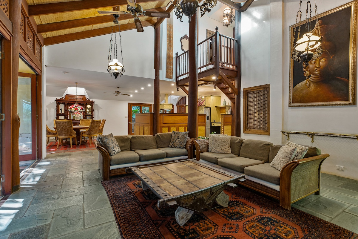 A spacious living area features an arrangement of comfortable seating, with a wooden coffee table at the center. High ceilings are complemented by wooden beams, while decorative lights and a large artwork add character. A dining area and kitchen are visible in the background.