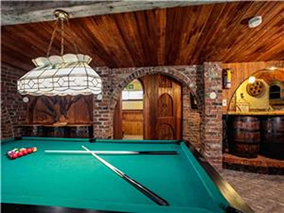 A games room features a green pool table, with cues laid across the surface. The space is accented by exposed brick walls and wooden beams, creating an inviting rustic atmosphere. An arched doorway leads to an adjoining area with barrel-style furniture.