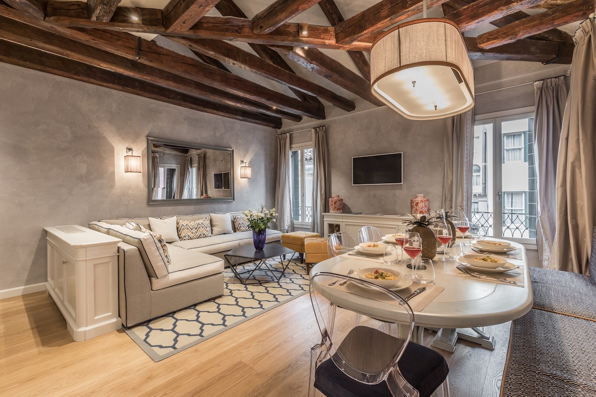 An open-plan living and dining area features tall windows with decorative balustrades, allowing natural light to fill the space. A modern kitchen is complemented by a stylish dining table set for four. Exposed wooden beams add character to the elegant, airy atmosphere.
