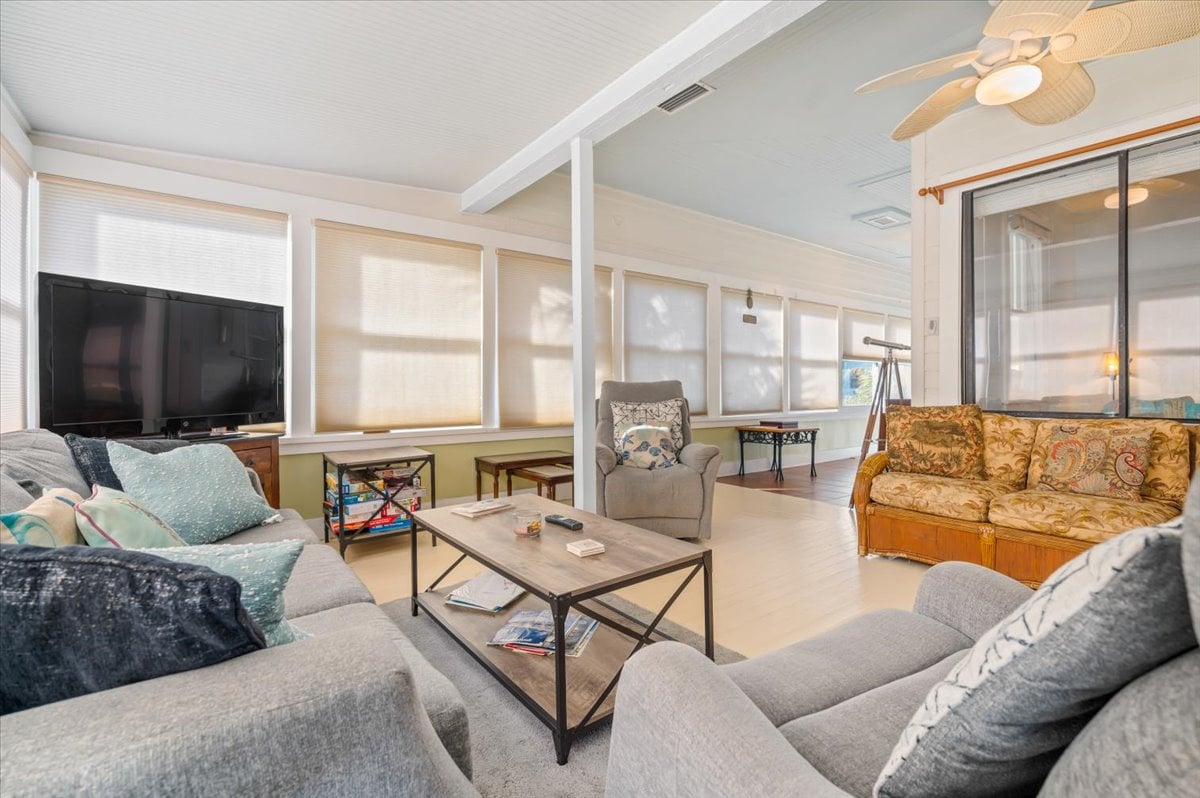 A bright and airy living area features large windows with sheer treatments, allowing ample natural light. Comfortable seating is arranged around a coffee table, with a television visible nearby. A small dining table stands in the background, providing an inviting space for gatherings.