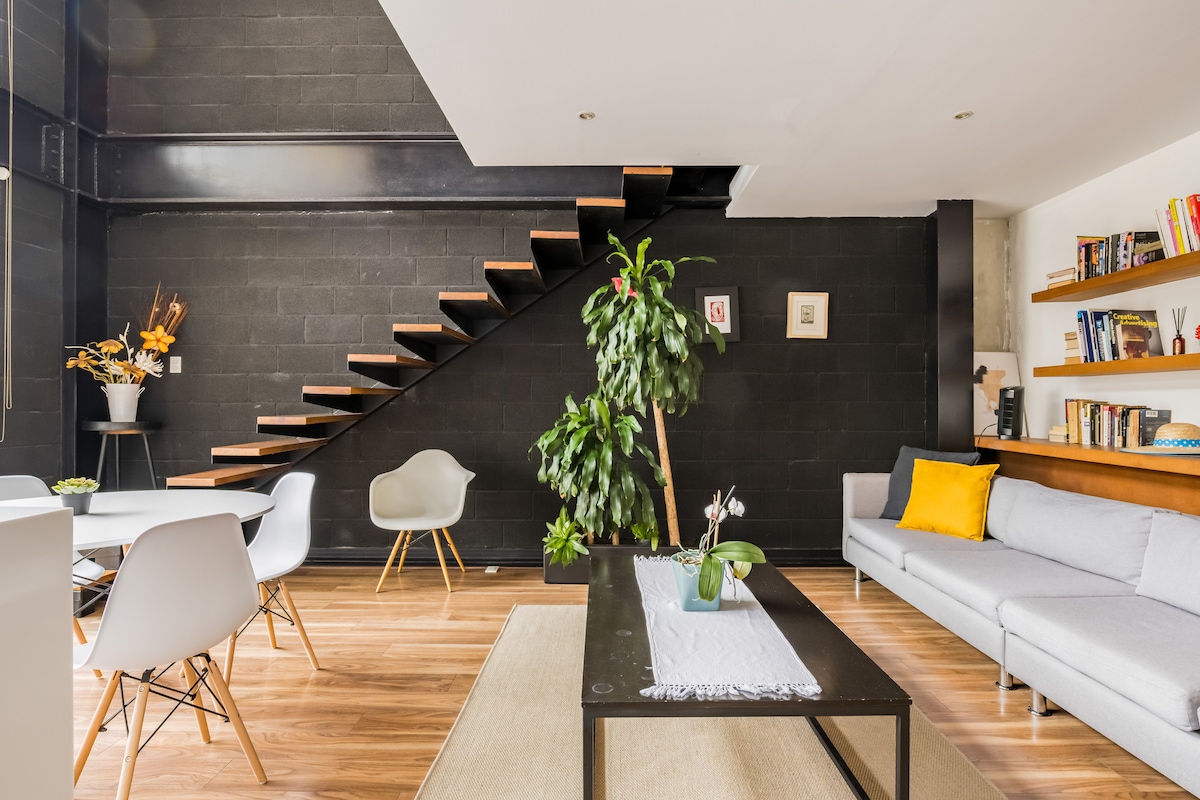 An open living area features a sleek design with warm wooden flooring. A modern gray sofa is placed adjacent to a simple coffee table, while a decorative houseplant adds a touch of greenery. A spiral staircase rises against a contrasting black wall, enhancing the space's contemporary feel.
