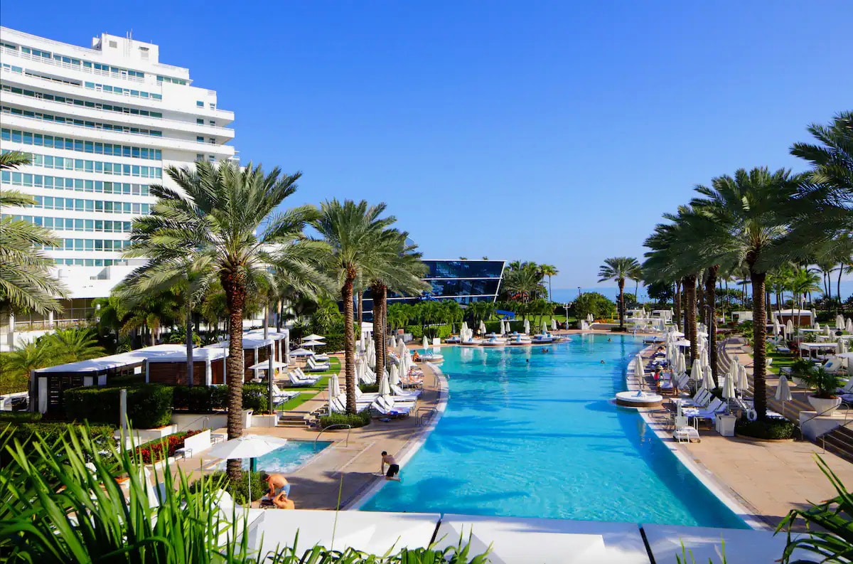 A large swimming pool extends through the resort, surrounded by tropical palm trees and lounge chairs. The sleek hotel buildings are visible in the background against a clear blue sky, creating a serene outdoor space for relaxation.